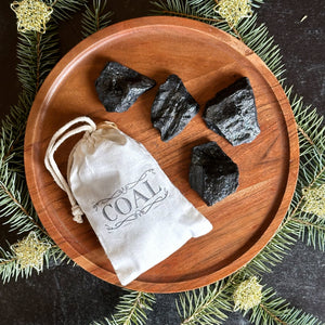A little muslin bag that has the word coal stamped on it is lying next to 4 pieces of black soap that are shaped like coal on a wooden plate. There are pine clippings arranged around the plate on a black background.